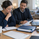 Two adults review documents together at a table beside an open laptop, notebooks and folders