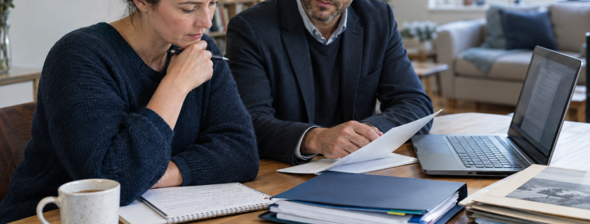 Two adults review documents together at a table beside an open laptop, notebooks and folders
