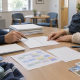 Two people review documents and a timetable at a table, with folders, stationery and a backpack nearby