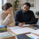Two people review documents at a desk while a third person works in the background