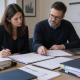 Two people reviewing and annotating documents at a desk with a laptop and organised files
